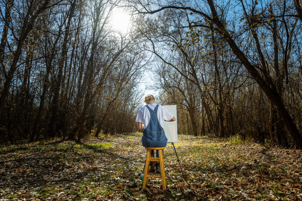 High school senior girl sitting in a grove of trees in Weston, Missouri, painting, captured by a top Kansas City senior photographer with a focus on natural beauty and serenity