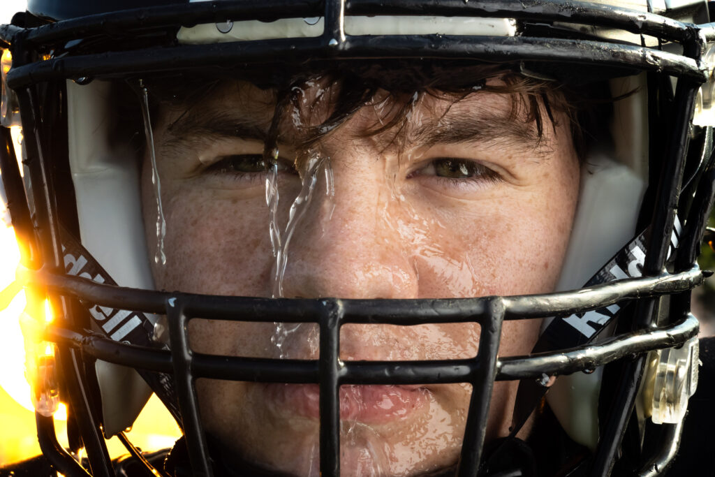 Up-close shot of a high school senior boy football player in a helmet with water pouring over him, featuring intense eyes, captured by a top Kansas City senior photographer