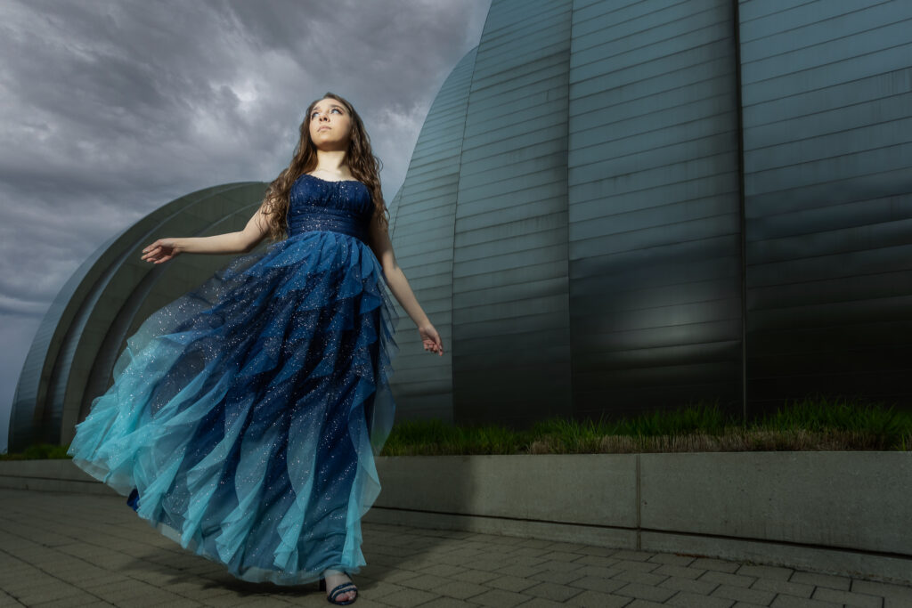 High school senior girl running in a beautiful gown in front of the Kaufmann Center for the Performing Arts with a dramatic sky and lighting, captured by the best Kansas City senior photographer