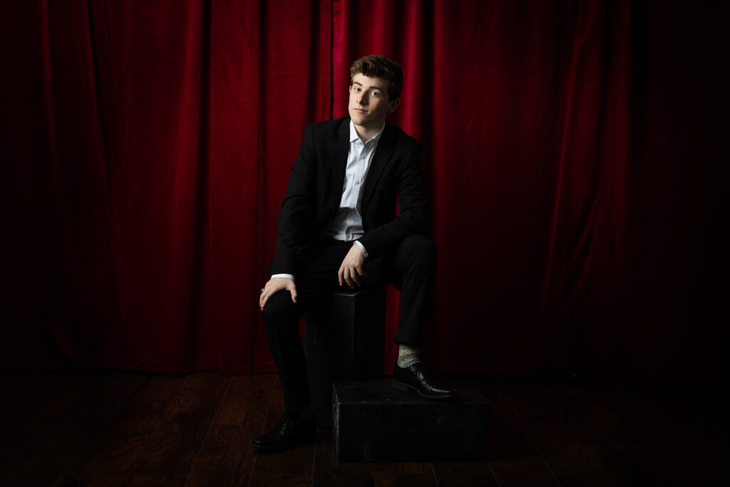 High school senior boy in a suit sitting in front of a red theater curtain, captured by a Kansas City senior photographer for a classic and polished portrait session.