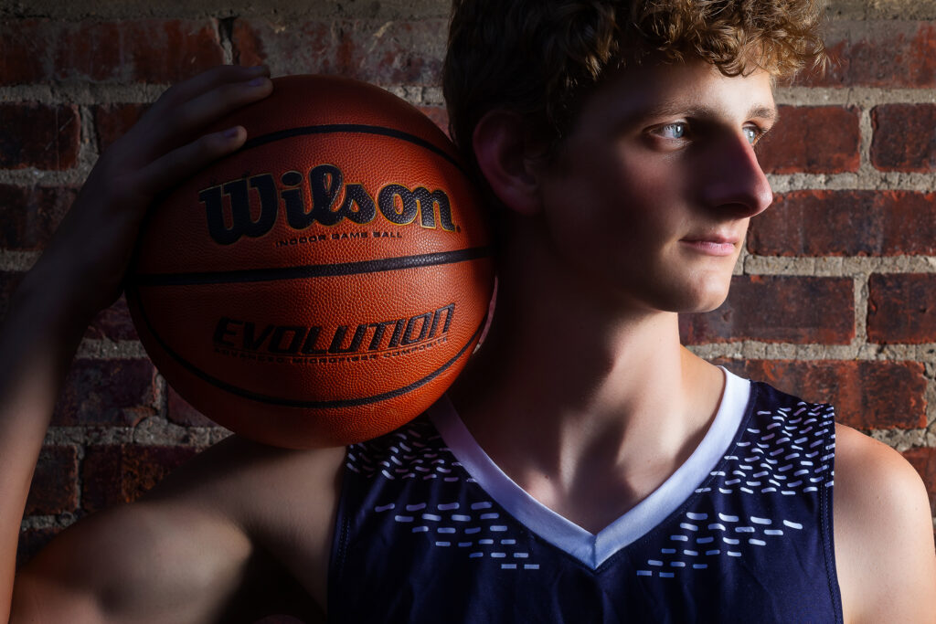 High school senior boy basketball player holding a basketball under dramatic lighting, captured by a Kansas City senior photographer for a powerful and athletic portrait session.